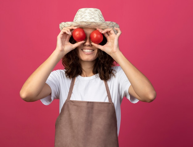 Jardinero mujer joven sonriente en uniforme con sombrero de jardinería mostrando gesto de mirada con tomates aislados en rosa