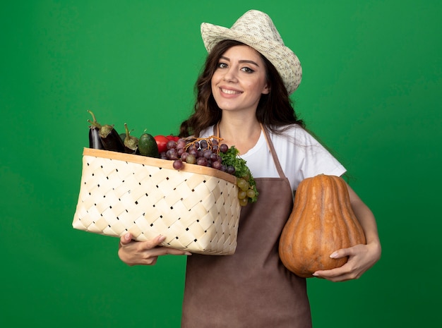 Foto gratuita jardinero mujer joven sonriente en uniforme con sombrero de jardinería tiene canasta de verduras y calabaza aislado en la pared verde con espacio de copia