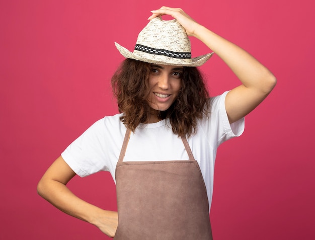 Jardinero mujer joven sonriente en uniforme vistiendo sombrero de jardinería poniendo la mano en la cadera sosteniendo el sombrero aislado en rosa