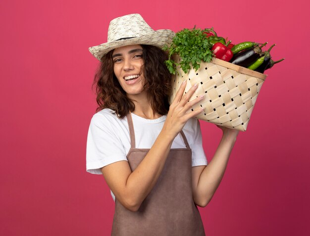 Jardinero mujer joven sonriente en uniforme vistiendo sombrero de jardinería sosteniendo canasta de verduras en el hombro aislado en rosa