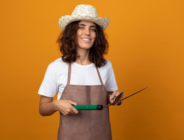 Jardinero mujer joven sonriente en uniforme vistiendo sombrero de jardinería sosteniendo tijeras