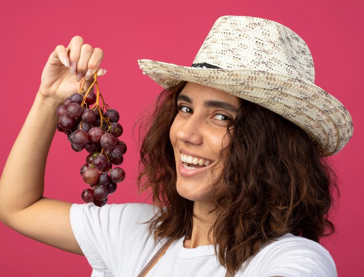 Foto gratuito jardinero mujer joven sonriente en uniforme vistiendo sombrero de jardinería sosteniendo uvas aisladas en rosa