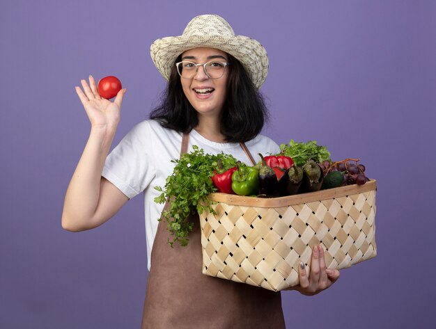 Jardinero mujer morena joven alegre en gafas ópticas y en uniforme con sombrero de jardinería sostiene cesta de verduras y tomate aislado en la pared púrpura