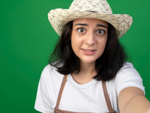 Foto gratuita jardinero mujer morena joven ansioso en gafas ópticas y uniforme con sombrero de jardinería sostiene y mira al frente aislado en la pared verde