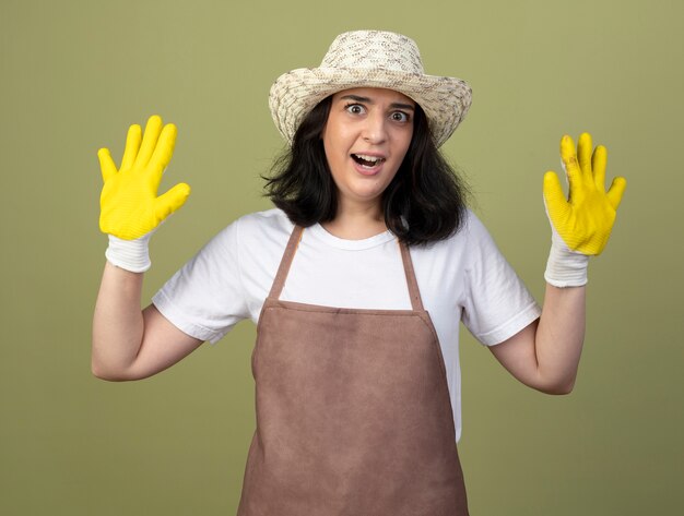 Jardinero mujer morena joven ansioso en uniforme con sombrero y guantes de jardinería se encuentra con las manos levantadas aisladas en la pared verde oliva