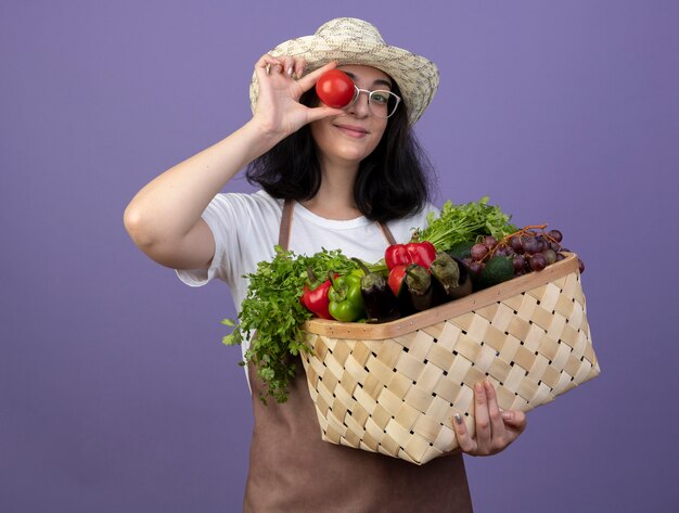 Jardinero mujer morena joven complacido con gafas ópticas y uniforme con sombrero de jardinería sostiene canasta de verduras y cubre el ojo con tomate aislado en la pared púrpura