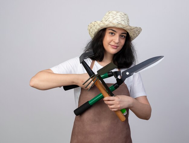 Jardinero mujer morena joven complacido en uniforme con sombrero de jardinería tiene herramientas de jardinería aisladas en la pared blanca con espacio de copia