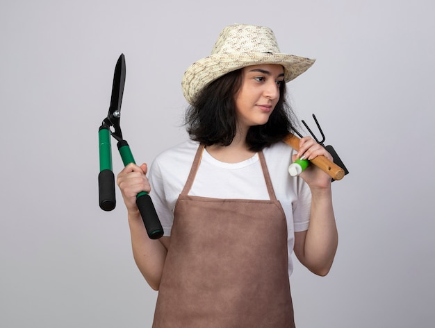 Jardinero mujer morena joven complacido en uniforme con sombrero de jardinería tiene herramientas de jardinería mirando al lado aislado en la pared blanca