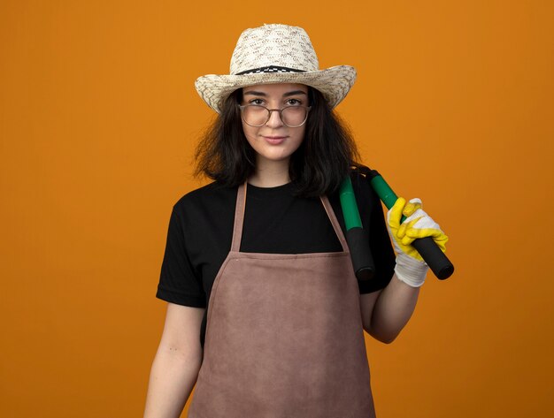 Jardinero mujer morena joven confiado en gafas ópticas y en uniforme con sombrero y guantes de jardinería sostiene podadoras de jardín en el hombro aislado en la pared naranja con espacio de copia