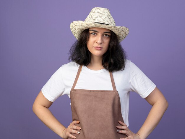 Jardinero mujer morena joven confiado en uniforme con sombrero de jardinería pone las manos en la cintura y mira al frente aislado en la pared púrpura
