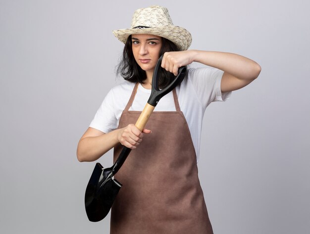 Jardinero mujer morena joven confiado en uniforme con sombrero de jardinería sostiene pala en blanco