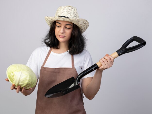 Jardinero mujer morena joven confiado en uniforme con sombrero de jardinería sostiene la pala y mira repollo aislado en la pared blanca con espacio de copia