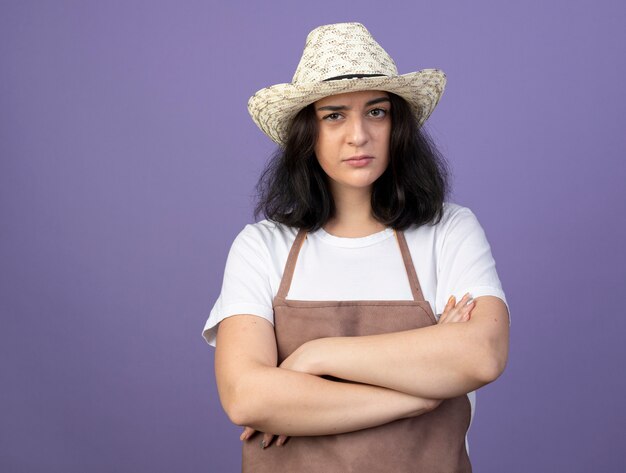 Jardinero mujer morena joven confidente en uniforme con sombrero de jardinería se encuentra con los brazos cruzados aislados en la pared púrpura