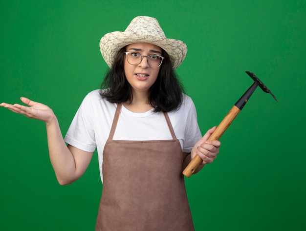 Jardinero mujer morena joven confundido con gafas ópticas y uniforme con sombrero de jardinería se encuentra con la mano levantada y sostiene rastrillo aislado en la pared verde