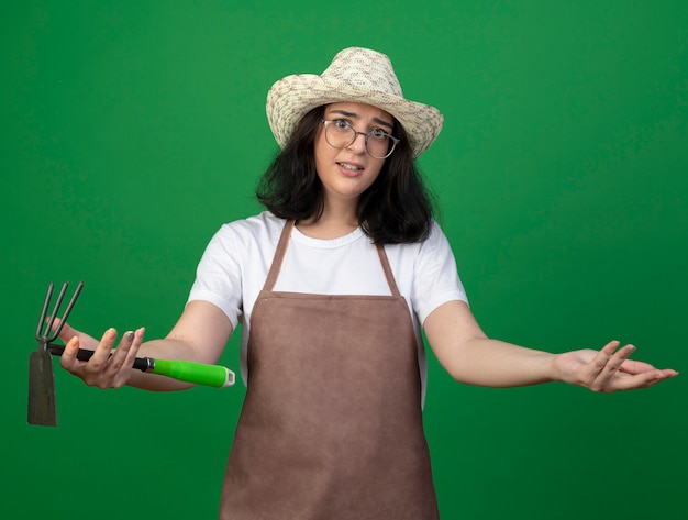 Jardinero mujer morena joven decepcionado con gafas ópticas y uniforme con sombrero de jardinería sostiene rastrillo de azada y mantiene la mano abierta aislada en la pared verde