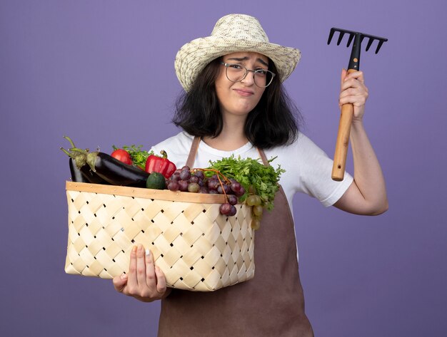 Jardinero mujer morena joven disgustado en gafas ópticas y en uniforme con sombrero de jardinería sostiene cesta de verduras y rastrillo aislado en la pared púrpura