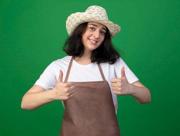 Jardinero mujer morena joven sonriente en gafas ópticas y uniforme con sombrero de jardinería pulgar hacia arriba de dos manos aisladas en la pared verde