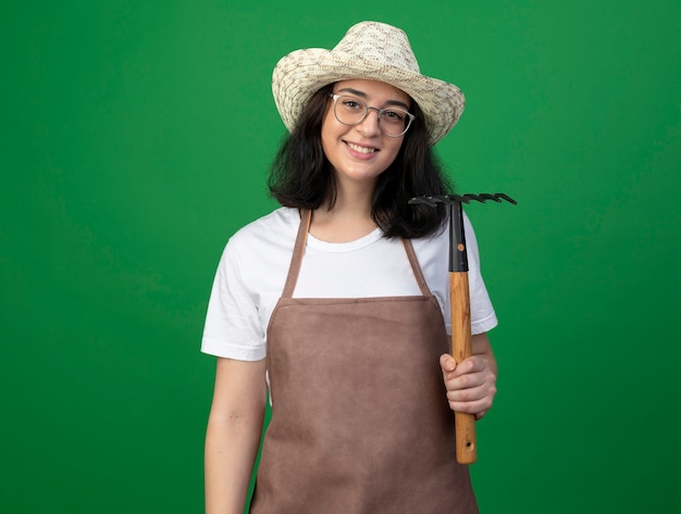 Foto gratuita jardinero mujer morena joven sonriente en gafas ópticas y uniforme con sombrero de jardinería sosteniendo rastrillo aislado en la pared verde