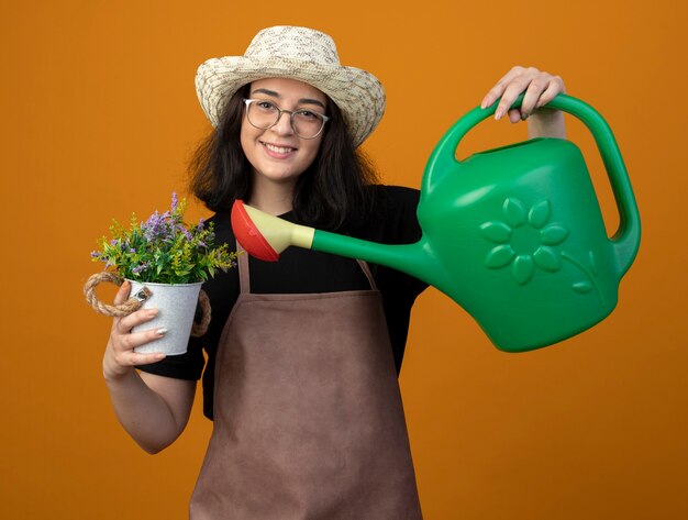 Jardinero mujer morena joven sonriente en gafas ópticas y en uniforme con sombrero de jardinería sosteniendo regadera sobre maceta aislada en pared naranja con espacio de copia
