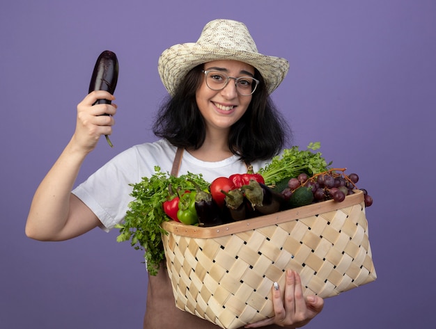 Jardinero mujer morena joven sonriente en gafas ópticas y en uniforme con sombrero de jardinería sostiene canasta de verduras y berenjena aislado en la pared púrpura