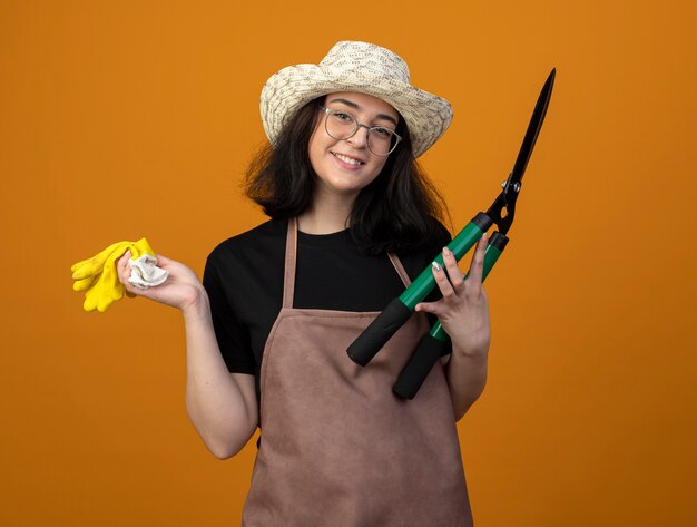 Jardinero mujer morena joven sonriente en gafas ópticas y en uniforme con sombrero de jardinería sostiene cortaúñas y guantes aislados en la pared naranja con espacio de copia