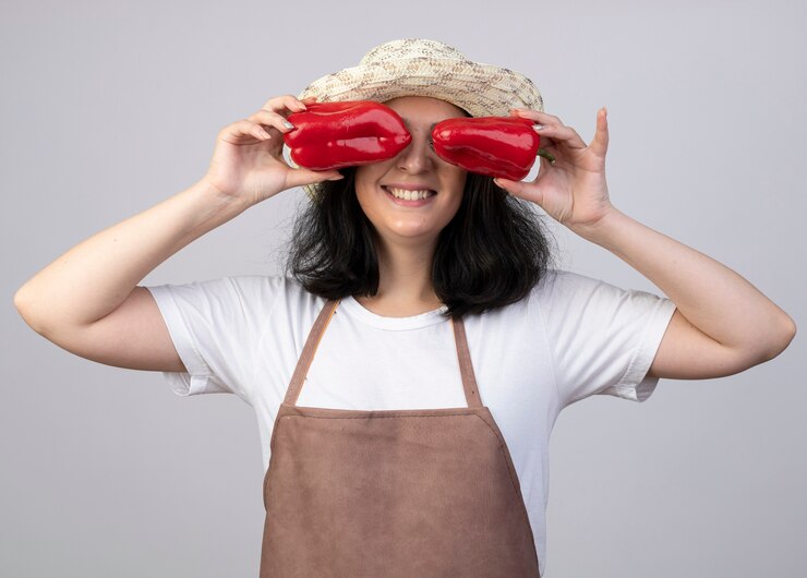 Foto gratuito jardinero mujer morena joven sonriente en uniforme con sombrero de jardinería cubre los ojos con pimientos rojos aislados en la pared blanca
