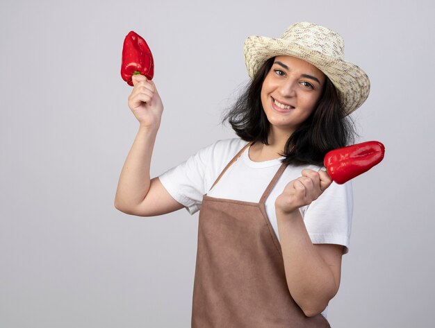 Jardinero mujer morena joven sonriente en uniforme con sombrero de jardinería tiene pimientos rojos aislados en la pared blanca con espacio de copia