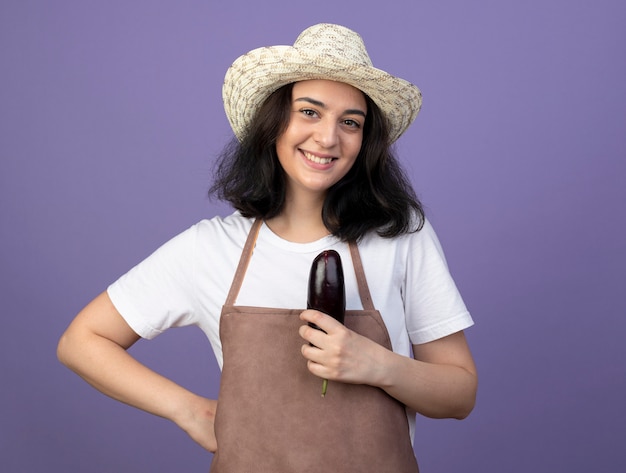 Jardinero mujer morena joven sonriente en uniforme vistiendo sombrero de jardinería sostiene berenjena aislado en la pared púrpura