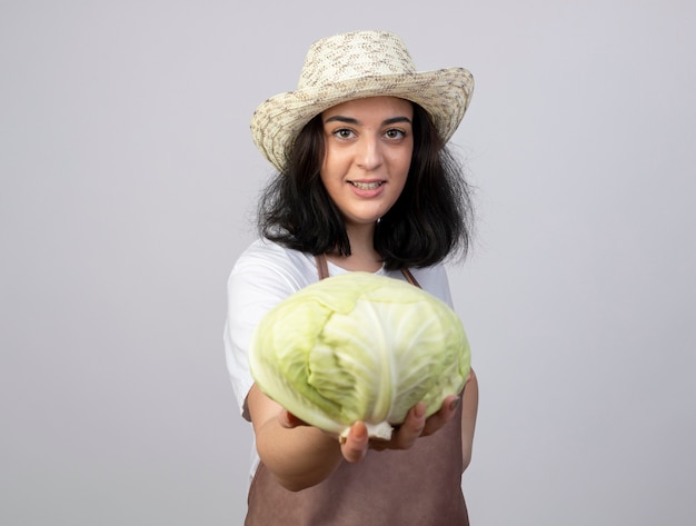 Jardinero mujer morena joven sonriente en uniforme vistiendo sombrero de jardinería sostiene repollo aislado en la pared blanca