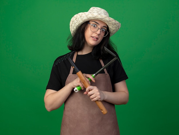 Foto gratuita jardinero mujer morena joven sorprendido con gafas ópticas y uniforme con sombrero de jardinería sostiene rastrillo de azada y rastrillo aislado en la pared verde