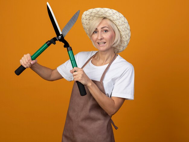 Jardinero rubio de mediana edad alegre mujer en uniforme con sombrero sosteniendo tijeras de podar aislado en la pared naranja con espacio de copia