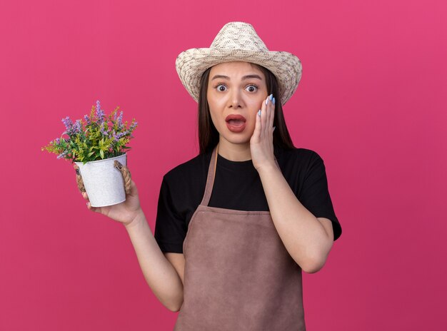 Jardinero de sexo femenino bastante caucásico ansioso que lleva el sombrero que cultiva un huerto pone la mano en la cara y sostiene la maceta en rosa