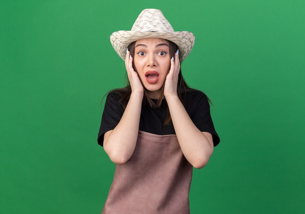Jardinero de sexo femenino bastante caucásico ansioso que lleva el sombrero que cultiva un huerto pone las manos en la cara aislada en la pared verde con el espacio de la copia