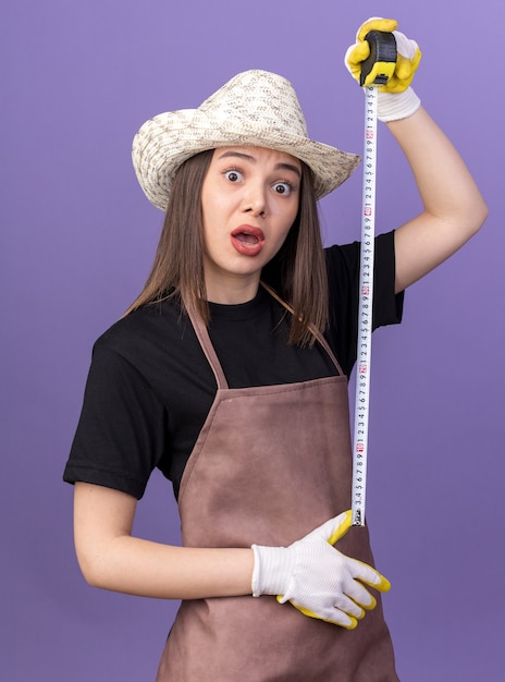 Jardinero de sexo femenino bastante caucásico ansioso con sombrero de jardinería y guantes sosteniendo cinta métrica aislada en la pared púrpura con espacio de copia