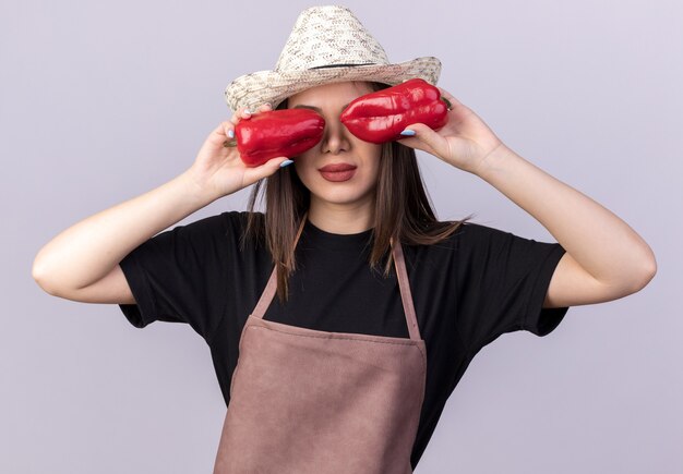 Jardinero de sexo femenino bastante caucásico confiado con sombrero de jardinería que cubre los ojos con pimientos rojos en blanco