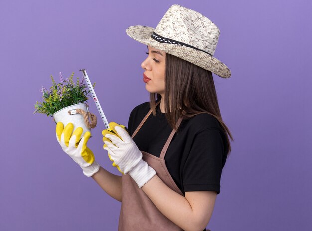 Foto gratuita jardinero de sexo femenino bastante caucásico seguro que lleva el sombrero de jardinería y guantes que miden la maceta con la cinta métrica en púrpura