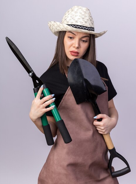 Jardinero de sexo femenino bastante caucásico seguro que lleva el sombrero de jardinería que sostiene la pala y que mira las tijeras de jardinería