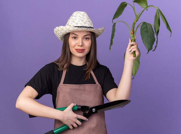 Jardinero de sexo femenino bastante caucásico seguro que lleva el sombrero de jardinería que sostiene las tijeras de jardinería y la rama de la planta en púrpura