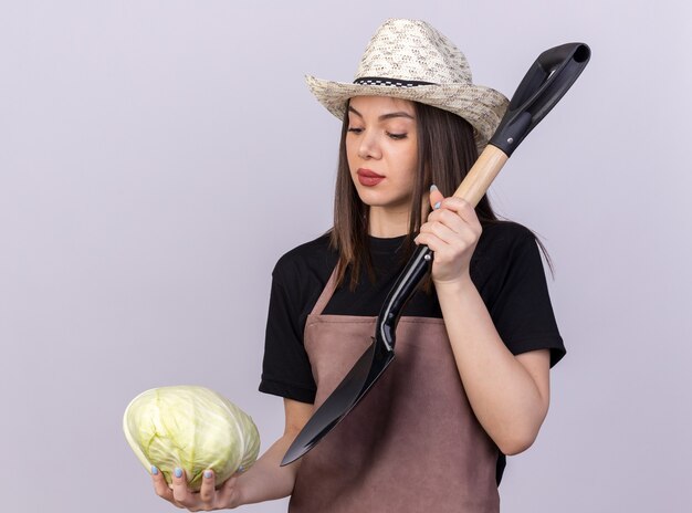 Jardinero de sexo femenino bastante caucásico seguro que lleva el sombrero que cultiva un huerto que sostiene la pala y que mira el repollo en blanco