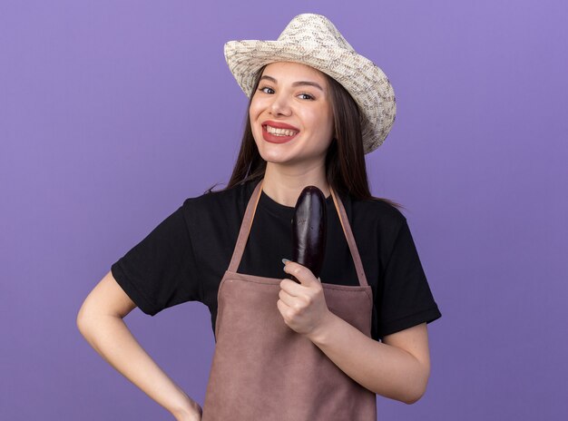 Jardinero de sexo femenino bastante caucásico sonriente que lleva el sombrero que cultiva un huerto que sostiene la berenjena en púrpura