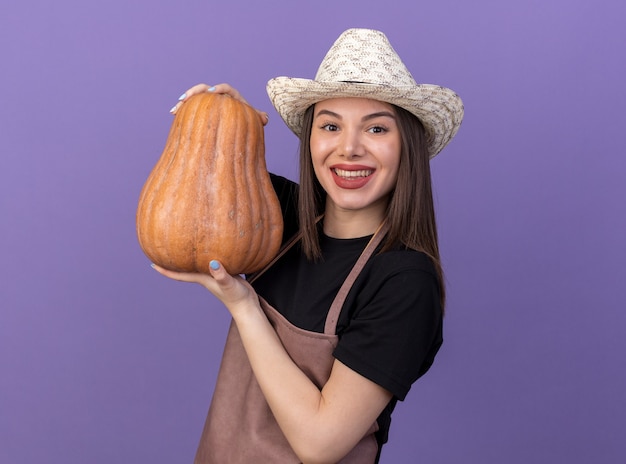 Jardinero de sexo femenino bastante caucásico sonriente que lleva el sombrero que cultiva un huerto que sostiene la calabaza aislada en la pared púrpura con el espacio de la copia