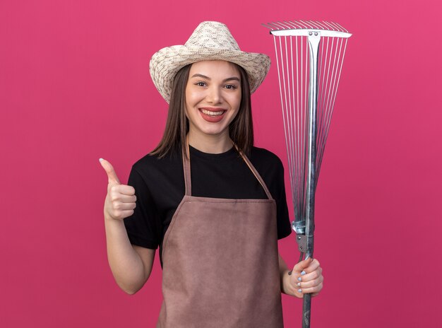 Jardinero de sexo femenino bastante caucásico sonriente que lleva el sombrero que cultiva un huerto que sostiene el rastrillo de la hoja y pulgar arriba en rosa