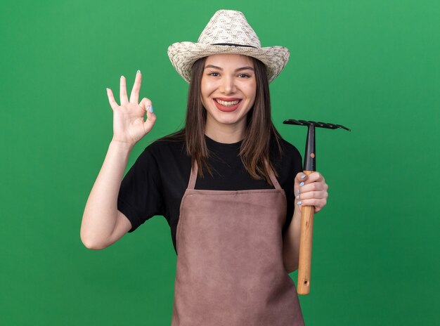 Jardinero de sexo femenino bastante caucásico sonriente que lleva el sombrero que cultiva un huerto que sostiene el rastrillo y que gesticula el signo de ok en verde