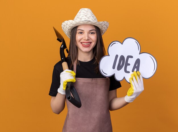 Jardinero de sexo femenino bastante caucásico sonriente con sombrero y guantes de jardinería sosteniendo la burbuja de la idea y la pala en el hombro aislado en la pared naranja con espacio de copia