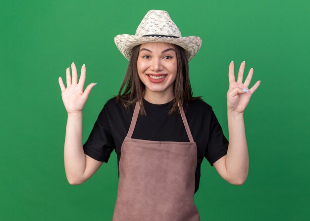 Jardinero de sexo femenino bastante caucásico sonriente con sombrero de jardinería gesticulando nueve con los dedos en verde