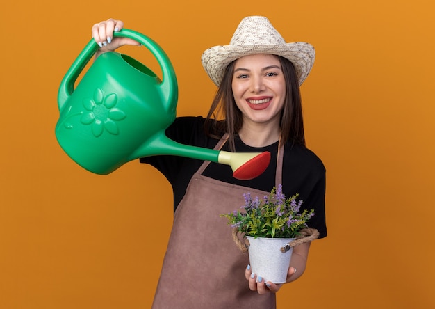 Jardinero de sexo femenino bastante caucásico sonriente con sombrero de jardinería pretende regar las flores en maceta con regadera aislado en la pared naranja con espacio de copia