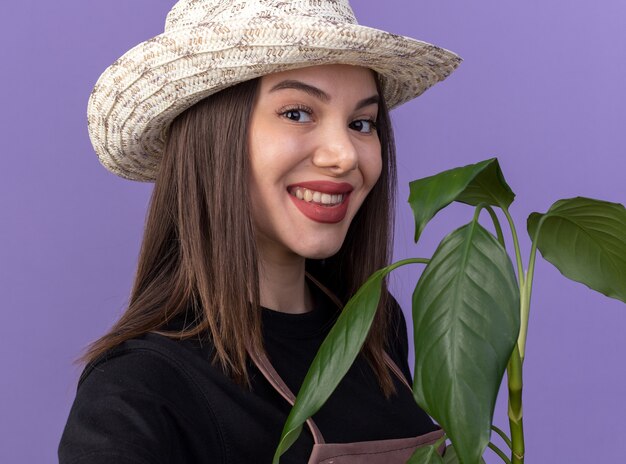 Jardinero de sexo femenino bastante caucásico sonriente con sombrero de jardinería sosteniendo la rama de la planta y aislado en la pared púrpura con espacio de copia