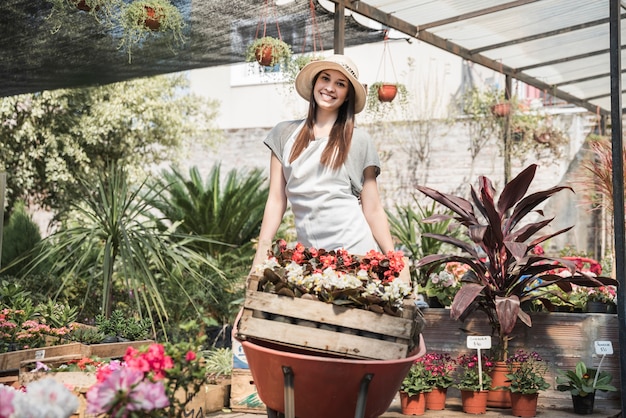 Jardinero de sexo femenino feliz que lleva el cajón de flores en carretilla