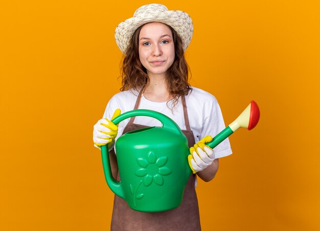 Jardinero de sexo femenino joven complacido con sombrero de jardinería con guantes sosteniendo regadera aislado en la pared naranja