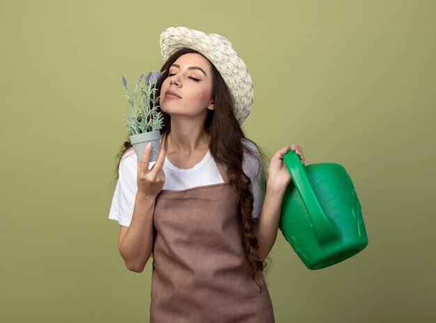 Jardinero de sexo femenino joven complacido en uniforme con sombrero de jardinería sostiene regadera y huele flores en maceta aislado en la pared verde oliva